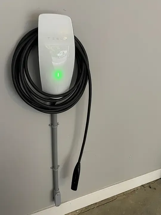 A wall-mounted EV charger with its cable coiled and a green ready light, conduit running down the wall